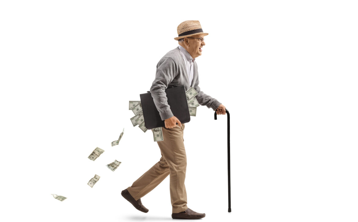Full length profile shot of an edlery man walking with a cane and carrying a briefcase full of money isolated on white background