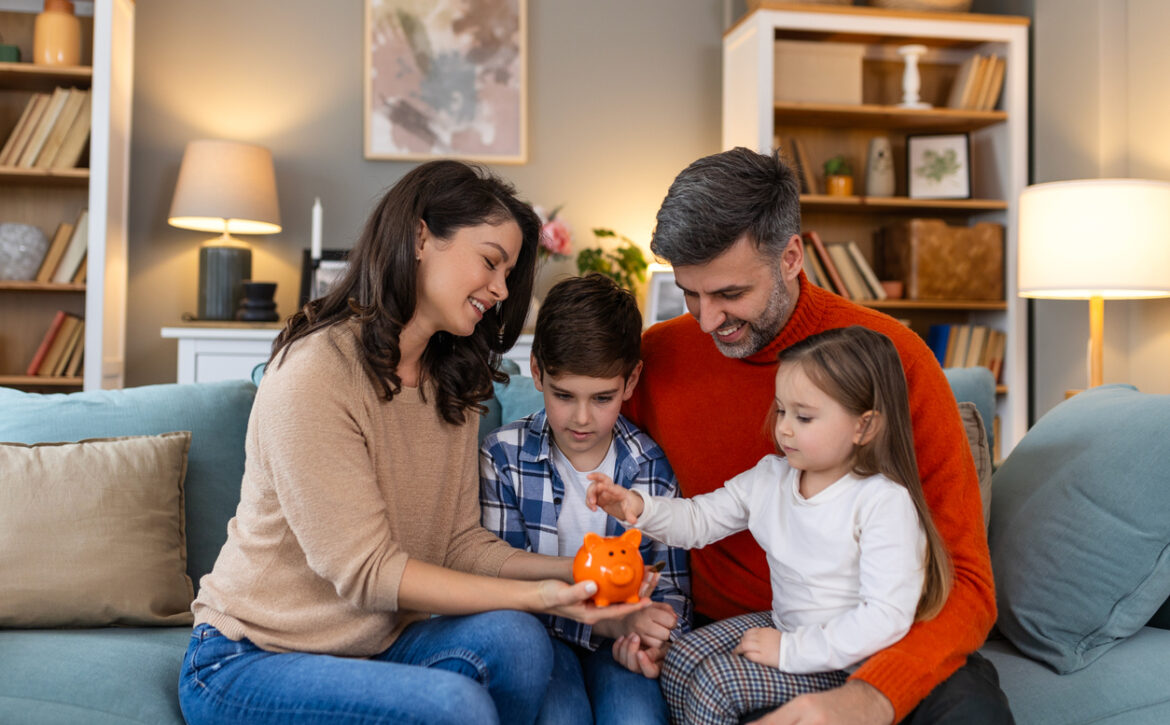 Happy family cheerful mother and father with kids smiling and putting coins into piggy bank while sitting on sofa at home