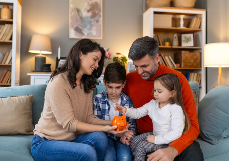 Happy family cheerful mother and father with kids smiling and putting coins into piggy bank while sitting on sofa at home